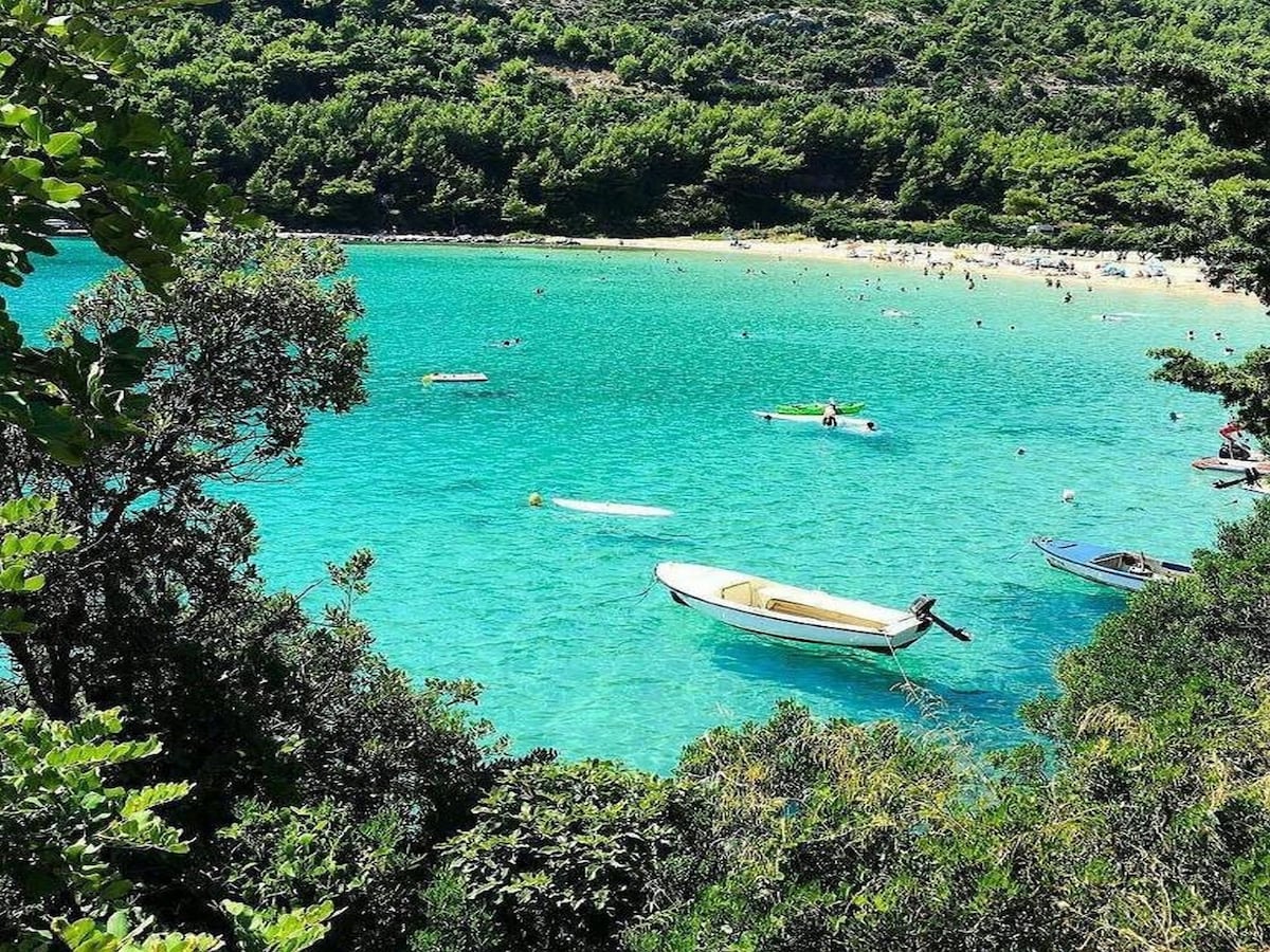 The image depicts a serene beach scene with clear turquoise waters and a white sandy shoreline. Small boats are anchored, and sunlight glistens on the water's surface. Lush greenery lines the beach, enhancing the natural beauty of the surroundings.