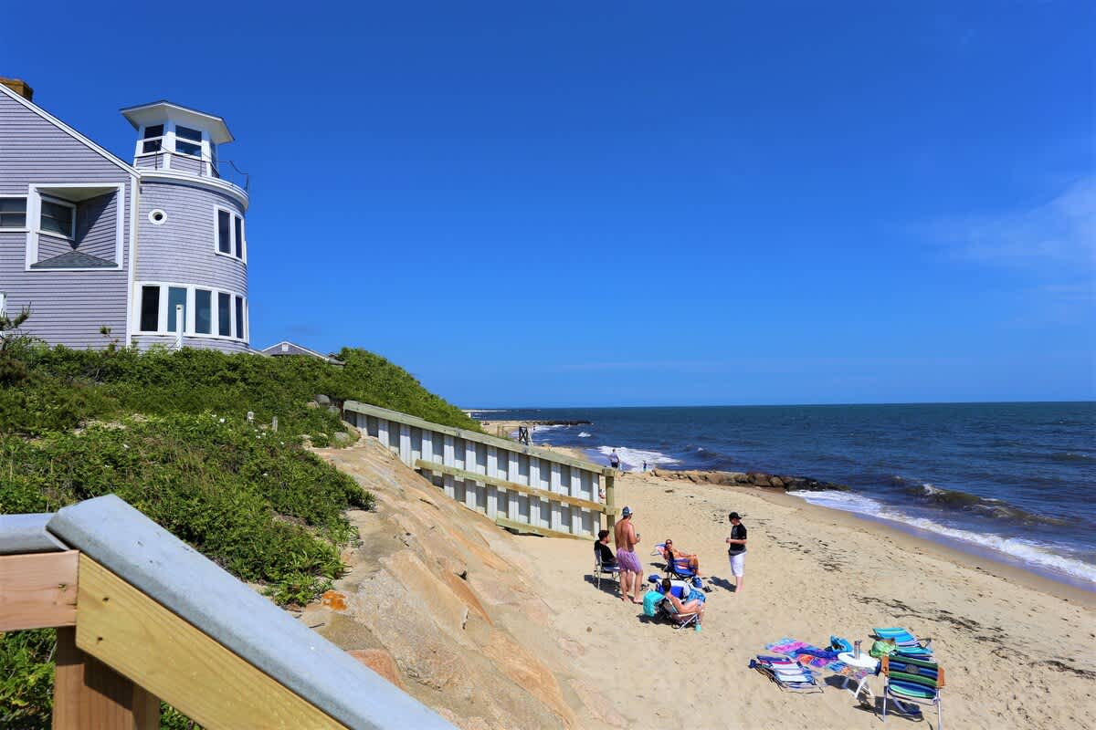 A sandy beach stretches along the shoreline, met by gentle waves under a clear blue sky. A group of beachgoers enjoys the sun near colorful beach chairs, while a building with a circular turret stands nearby, framing the picturesque coastal scene.