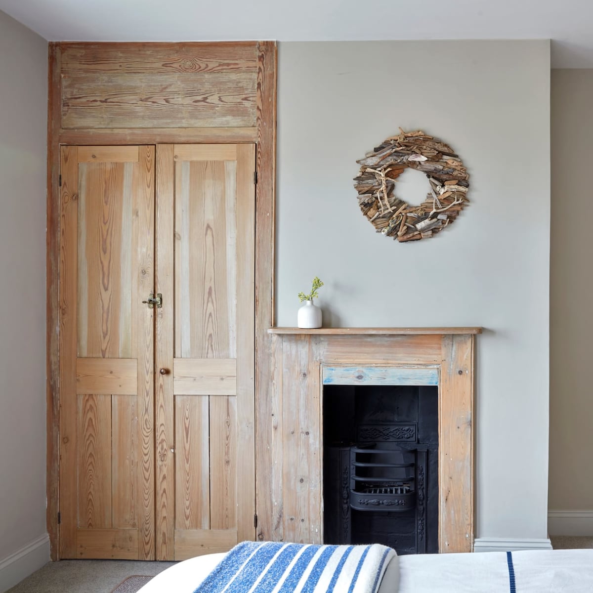 Natural wood finishes create a warm ambiance in the cozy bedroom. A simple, elegant wreath made of twigs adorns the wall above a rustic wooden fireplace, which features a classic black insert. Light-colored walls enhance the serene, calming environment.