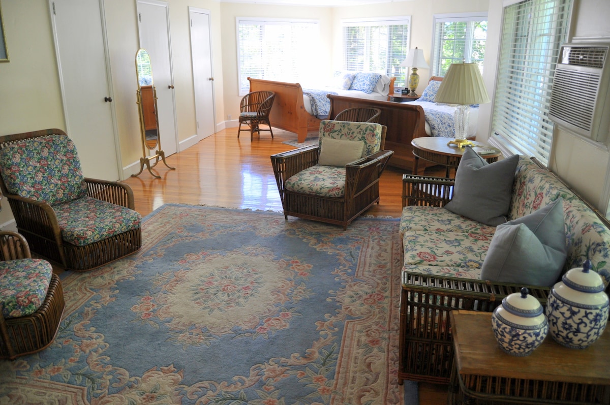 A cozy sitting area features wicker furniture with floral upholstery, arranged around a decorative area rug. Large windows allow natural light to fill the space, revealing a view into the guest room with a spacious bed and additional furnishings in the background.