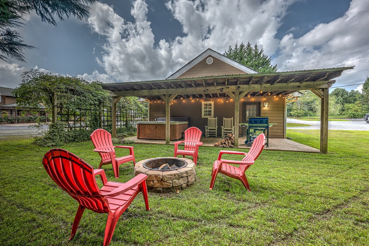 A cozy outdoor space features a circular arrangement of five bright red Adirondack chairs surrounding a stone fire pit. A wooden structure with a roof provides shade, alongside a hot tub and a seating area against the backdrop of a grassy yard.