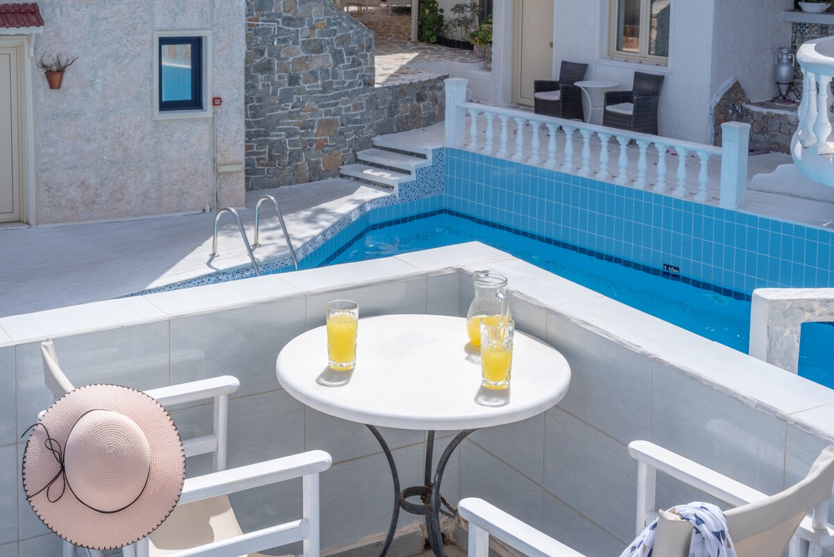 A patio overlooks a refreshing pool area, featuring a small round table set with two glasses of juice and a pitcher. White chairs provide comfortable seating, while a wide-brimmed hat rests casually on the table, inviting relaxation by the water.