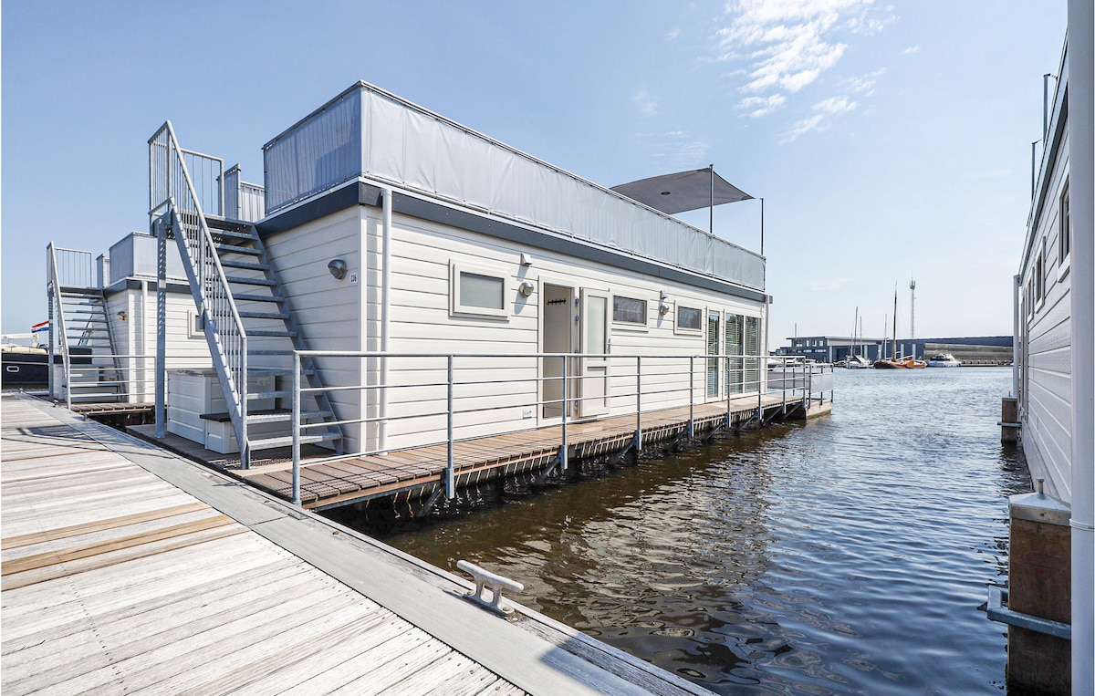 A modern floating vacation home is positioned along the water's edge, with large windows reflecting the sunlight. A staircase leads to a spacious roof terrace that overlooks the marina. The exterior features a light-colored finish, blending harmoniously with the surrounding environment.