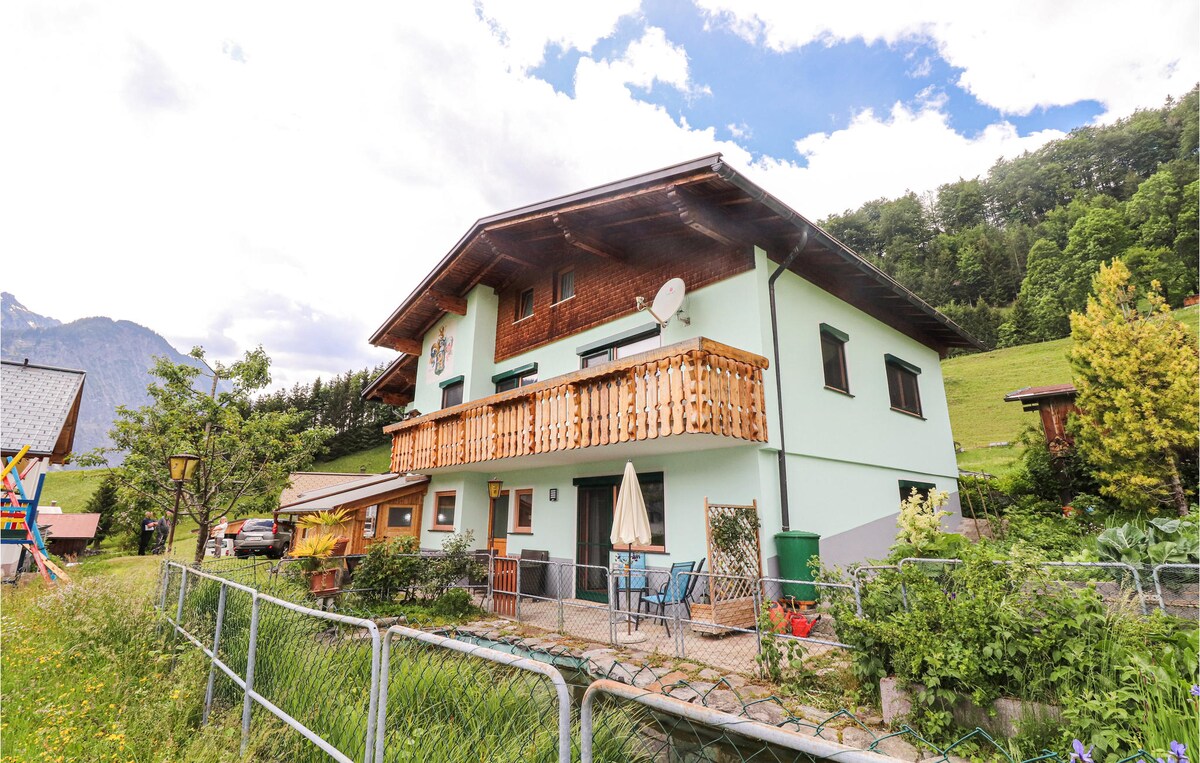 The two-story building exhibits a traditional alpine design with wooden accents. A balcony extends from the upper level, while a patio area featuring outdoor seating is accessible on the ground level. Lush greenery and mountains can be seen in the background.