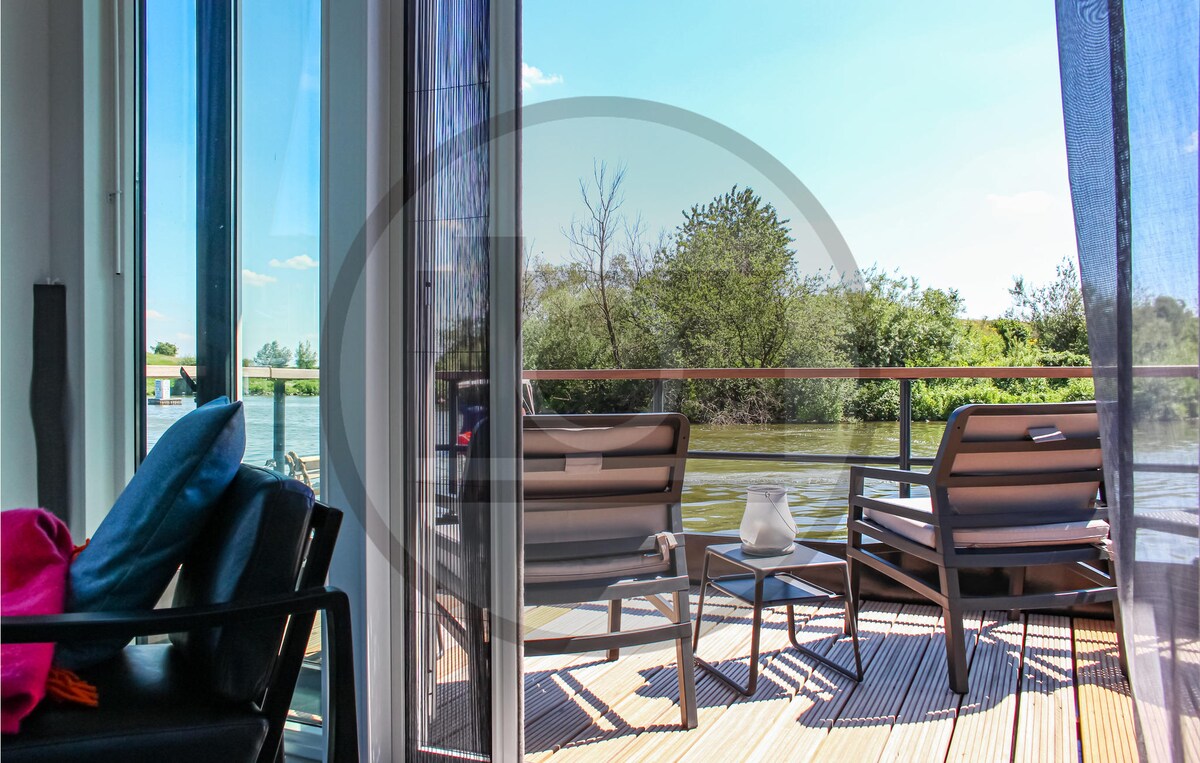 A serene terrace features two comfortable lounge chairs facing the water, surrounded by nature. The glass doors leading to the living area provide an unobstructed view of the tranquil surroundings, creating a peaceful outdoor space.