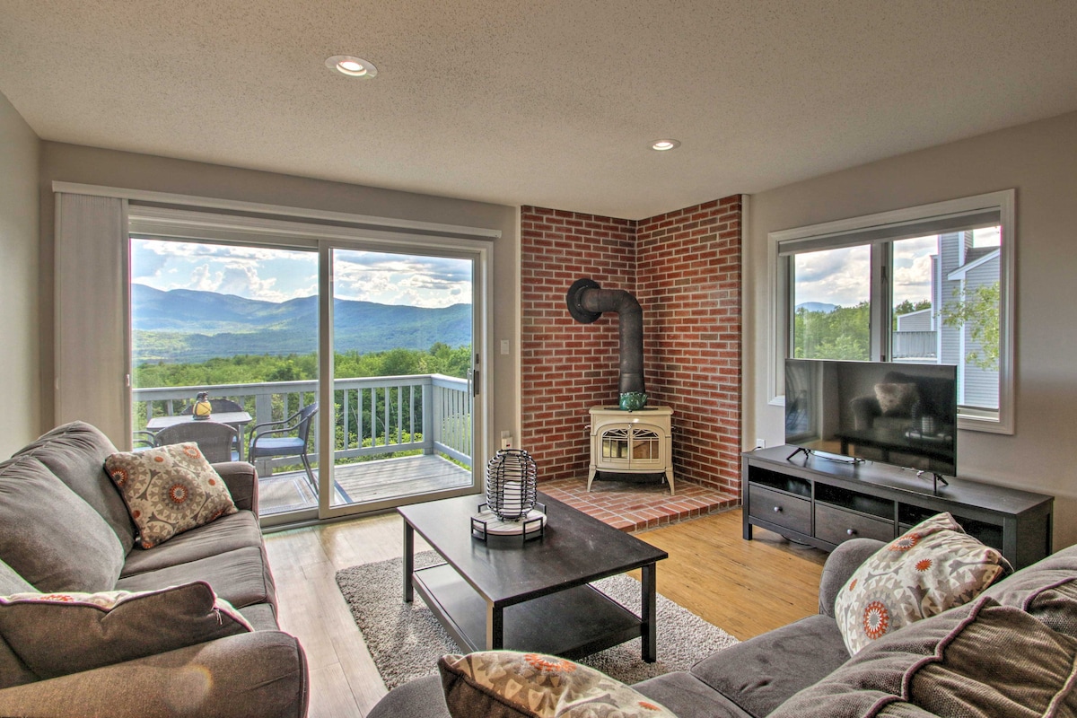 A comfortable living area features plush seating arranged around a central coffee table. Natural light floods the space through large sliding glass doors, which open onto a private balcony overlooking mountain vistas. A brick accent wall complements the propane gas stove, adding to the inviting ambiance.