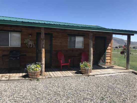 A rustic exterior of Cowboy Bunkhouse #2 is showcased, featuring a covered front porch with two red chairs and potted flowers. The entrance is flanked by wooden posts, and a gas grill is visible on the porch. Gravel leads to the entrance, surrounded by open landscapes.