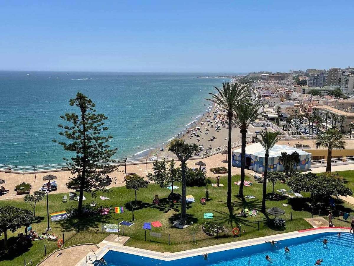 A panoramic view captures the beachfront and the shimmering sea, with golden sand and scattered beachgoers. Lush green gardens are visible in the foreground surrounding a gleaming pool, while palm trees sway gently in the warm breeze, enhancing the coastal atmosphere.
