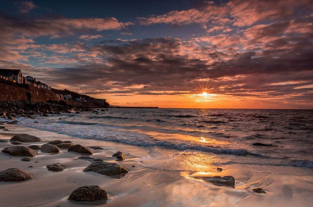The image captures a serene coastal scene featuring gentle waves lapping at a sandy shore, with smooth rocks scattered along the beach. A vibrant sunset casts warm hues across the sky, while buildings line the distant coastline against a backdrop of dramatic clouds.