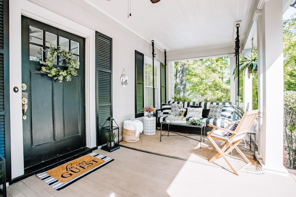 The front porch features a welcoming entry with a black door and decorative mat. A stylish seating arrangement includes striped cushions, complemented by a cozy atmosphere. Natural light filters in, while greenery decor and a rug enhance the inviting space.