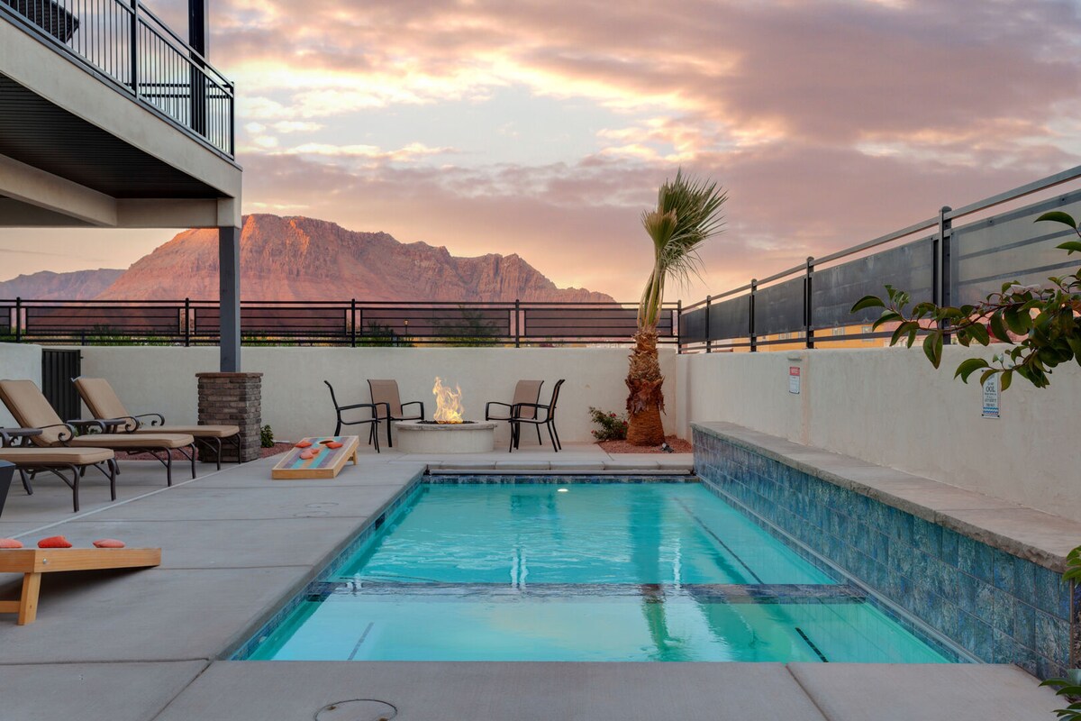 A private pool is shown in the foreground, featuring clear water under a sunset sky. Comfortable lounge chairs line the poolside, while a fire pit is positioned nearby, creating a peaceful ambiance. Mountains are visible in the background, adding a scenic backdrop.