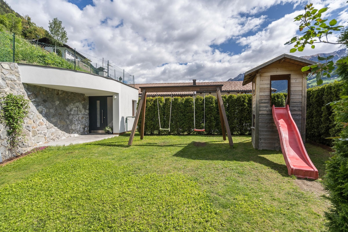 A green outdoor space is depicted, featuring a small playground with a wooden swing set and a slide. The yard is bordered by tall hedges, providing a sense of privacy. A stone wall is visible in the background, complementing the natural surroundings.