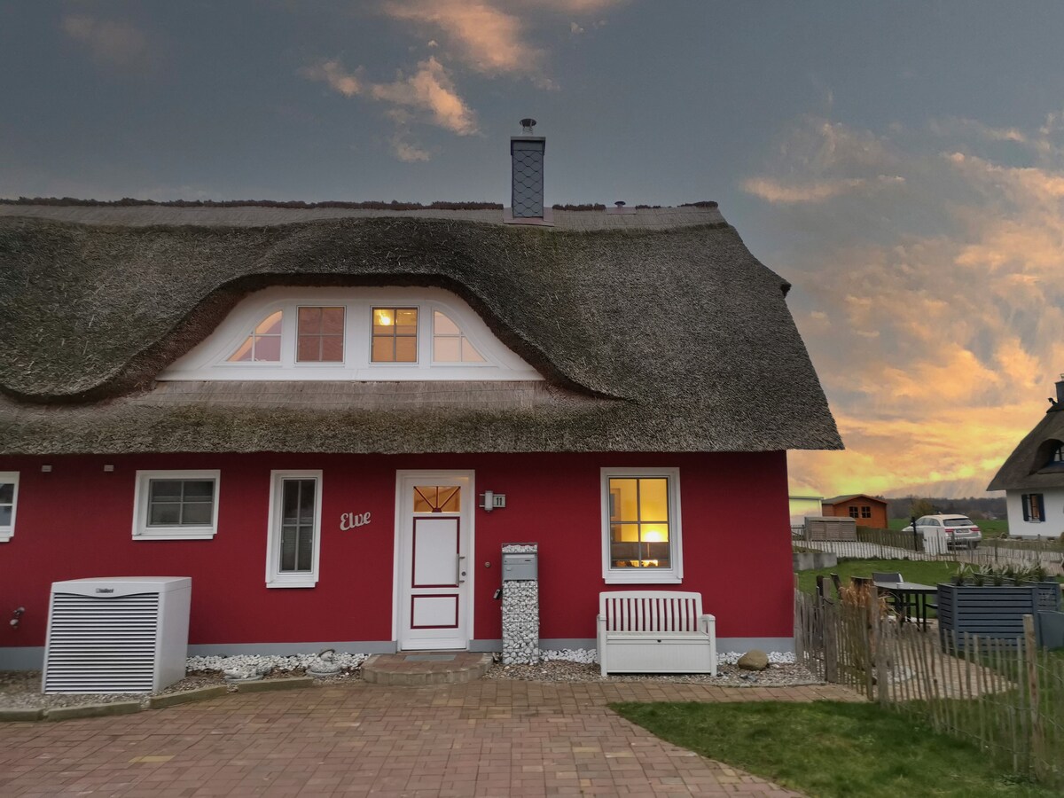 The thatched-roof house, painted in a warm red hue, stands prominently against a colorful sky at dusk. Windows reflect soft natural light, and a cozy outdoor area is visible, featuring stone pathways and a bench. A fenced boundary ensures privacy and security.