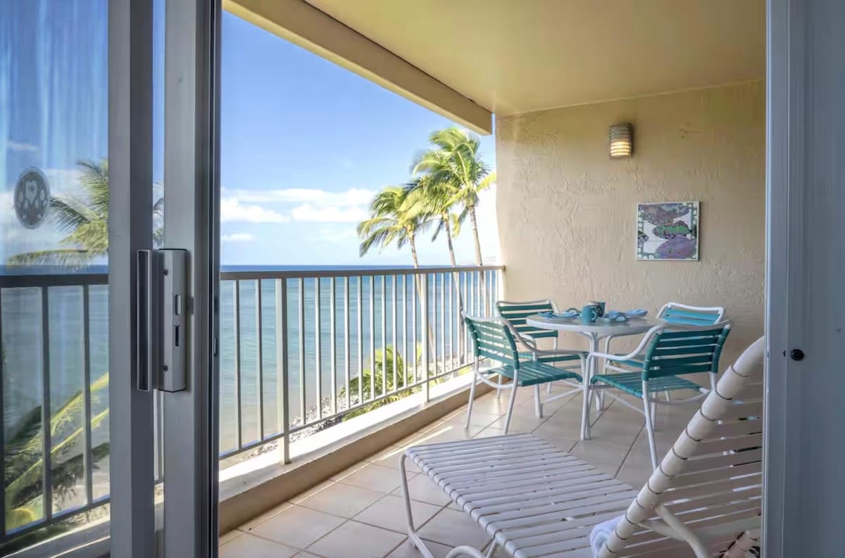 A private lanai offers ocean views and a light breeze, furnished with a round table and four chairs, alongside a lounge chair. Lush palm trees frame the scene, and the clear blue sky enhances the serene atmosphere of beachfront living.
