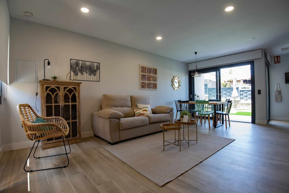 The open-plan living area features a spacious beige sofa positioned alongside a stylish wooden cabinet. A round table with chairs occupies the dining area, while large glass doors provide access to the outdoor space, allowing natural light to fill the room.