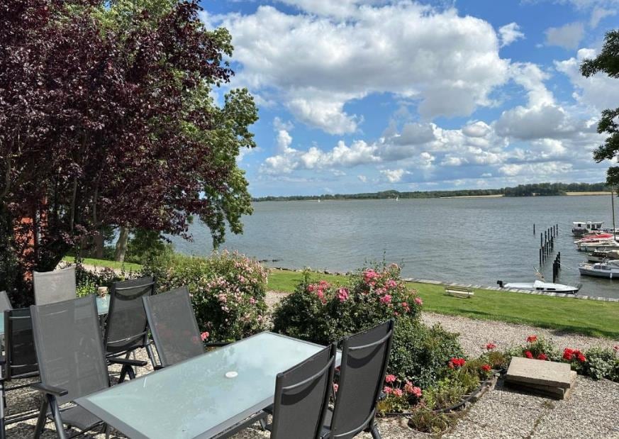 An outdoor terrace area features a glass table surrounded by six chairs. Lush greenery and colorful flower beds border the space, offering serene views of the water and distant shoreline under a partly cloudy sky.
