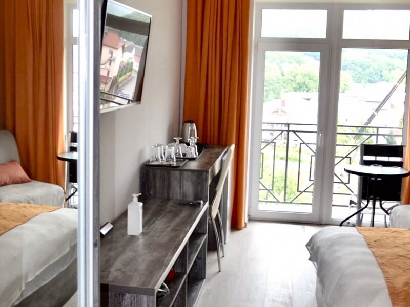 A modern room features two spacious beds and a comfortable armchair. A long wooden desk holds a kettle and glasses. Large windows with orange curtains allow natural light to illuminate the room, while offering views of the surrounding landscape and balcony access.