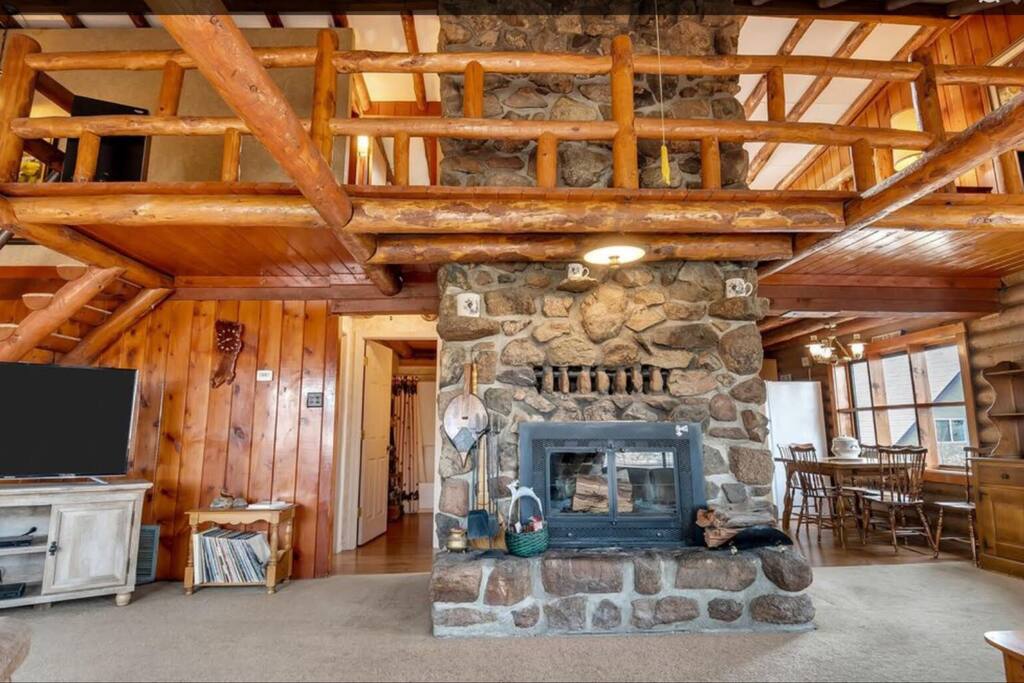 A spacious living area is highlighted by a large stone fireplace, surrounded by wooden beams and warm wood accents. A dining table with chairs is visible in the background, with soft natural lighting illuminating the space.