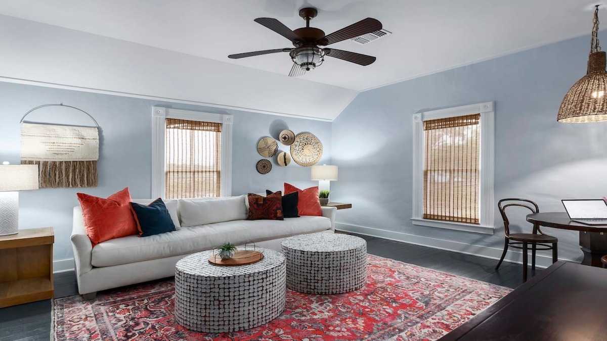 A light-filled living area is furnished with a comfortable white sofa adorned with colorful pillows. Two woven coffee tables sit at the center, surrounded by hardwood flooring and walls in soft blue. Natural light filters through bamboo shades on the windows, creating a serene environment.