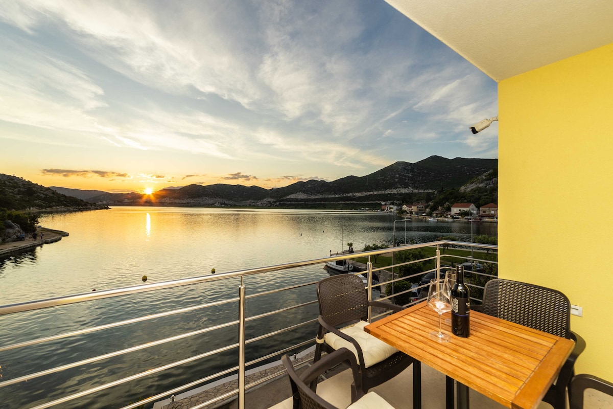 A furnished terrace overlooks the tranquil waters, with a table set for outdoor dining. Scenic views of the sunset reflect on the calm surface, while surrounding hills are visible in the background. Comfortable chairs complement the inviting atmosphere.