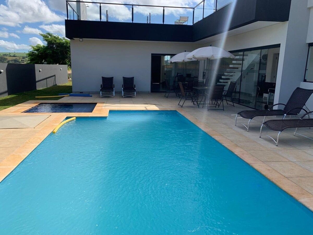 A bright outdoor area features a sparkling pool surrounded by tiles. Sun loungers are positioned on the deck, with umbrellas providing shade. A staircase leads to a terrace, where additional seating is visible, enhancing the space for relaxation and leisure.
