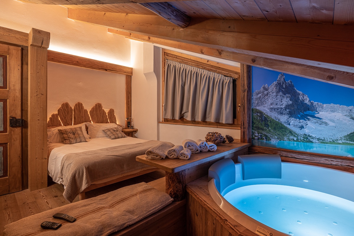 A relaxing bedroom features a comfortable double bed with a wooden headboard. Nearby, a modern hydromassage tub is illuminated softly, complementing the rustic wooden accents. Large windows display scenic mountain views, while warm lighting enhances the calming atmosphere.