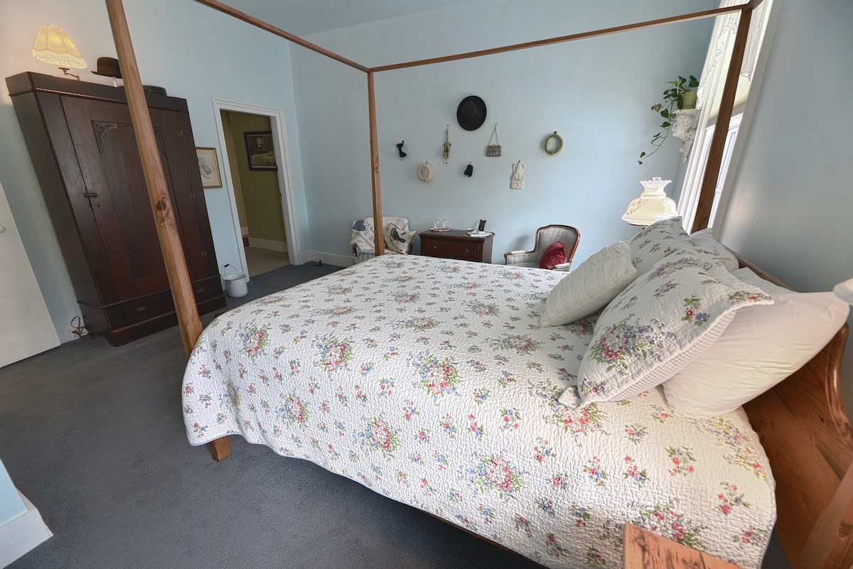 A spacious bedroom features a four-poster bed adorned with a floral quilt. Light blue walls complement a cozy reading nook and an antique dresser, creating a calming atmosphere. Natural light filters through a nearby window, casting soft shadows across the carpeted floor.