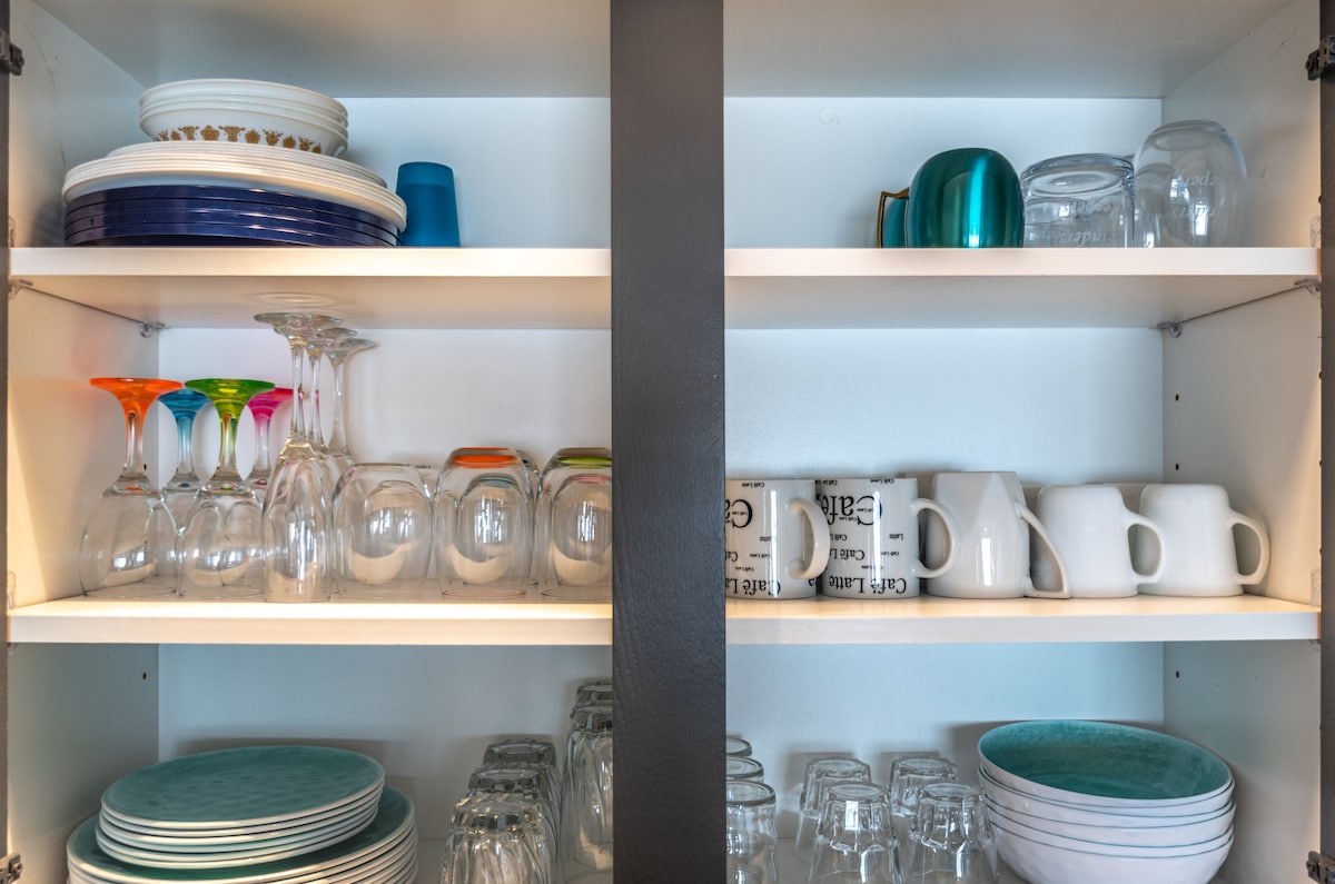 The kitchen cabinet displays an organized collection of dinnerware and glassware. Colorful wine glasses and clear tumblers sit atop, while ceramic plates and mugs are arranged neatly on the shelves below. Various glass containers are visible, providing a variety of dining options.
