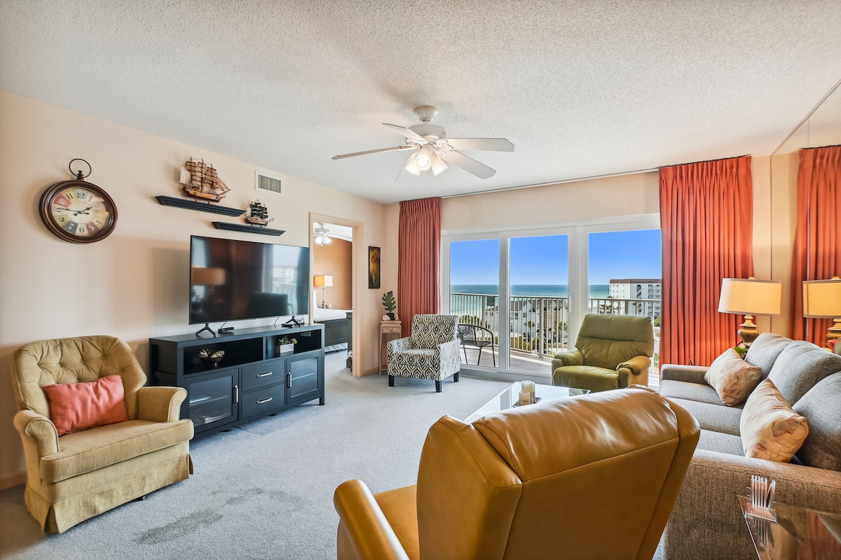 A spacious living area is presented with large windows offering a view of the outdoors. Comfortable seating includes a brown leather armchair, a light green chair, and a gray sofa. A television is mounted on the wall, and vibrant orange curtains frame the windows, complementing the decor.
