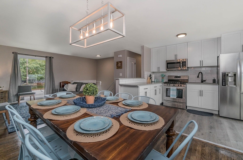 A modern kitchen with stainless steel appliances and white cabinetry is complemented by a spacious dining area featuring a dark wooden table. Plates and woven chargers are neatly arranged for eight, and a potted plant adds a touch of greenery. Natural light filters in through a nearby window.