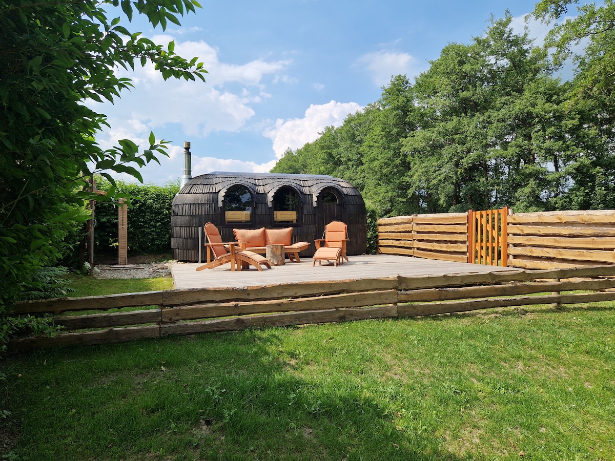 An outdoor terrace features garden furniture arranged under a clear sky, surrounded by greenery. The unique Igluhut, crafted from natural materials, showcases a distinctive design with large windows. A wooden fence outlines the area, providing a sense of privacy and connection to nature.