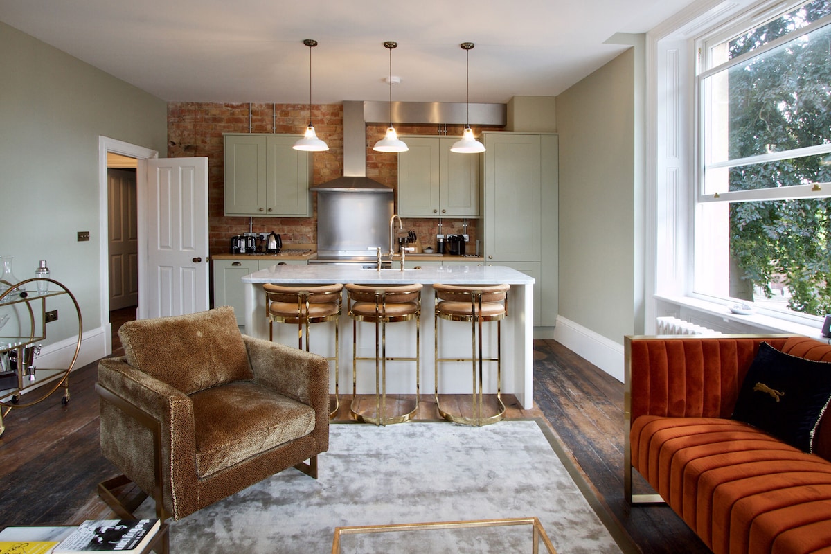 The open-plan kitchen and living area features a modern design with a large kitchen island surrounded by three bar stools. Natural light fills the space through large windows, highlighting the warm tones of the furnishings, including a cozy armchair and a plush sofa.
