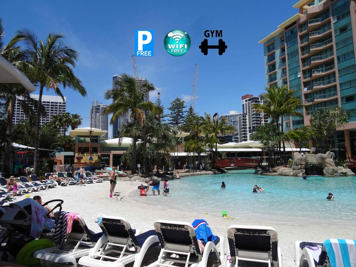 A resort-style pool area features a sandy beach-like entry, surrounded by palm trees and lounge chairs. Guests enjoy the water, with a backdrop of the city skyline. The pool is complemented by tropical landscaping and an inviting atmosphere for relaxation and leisure.