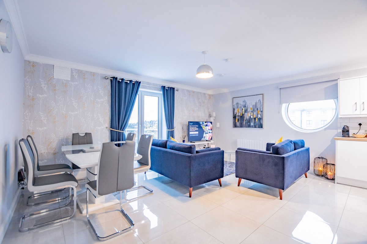An open-plan living and dining area features two velour sofas in deep blue, paired with a glass dining table that seats six. Natural light floods through the window, illuminating a modern kitchen in the corner. Stylish decor elements enhance the contemporary setting.
