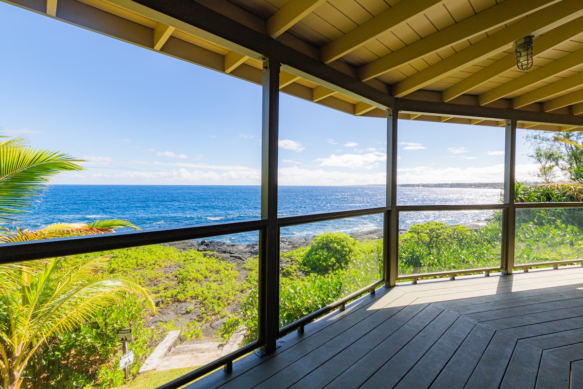 A covered lanai offers a panoramic view of the sapphire blue ocean and green coastal flora. Large windows allow natural light to illuminate the space, with a wooden floor providing a warm and inviting feel. The tranquil atmosphere is enhanced by the sounds of the ocean waves.