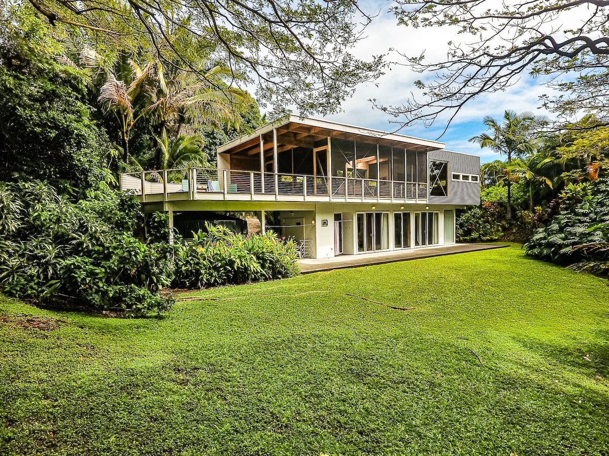 A modern house blends into its lush surroundings, featuring a spacious deck supported by a concrete foundation. Large glass doors provide a view of the green lawn and tropical foliage, while the roof extends to create a shaded area for outdoor relaxation.