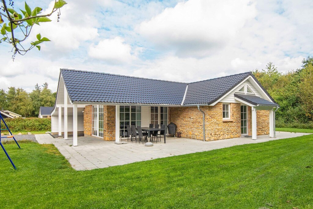 A spacious holiday home is set on a lush green lawn, featuring a large patio area with multiple chairs and a table. The exterior showcases a stone façade, complemented by a tiled roof and large windows providing ample natural light.
