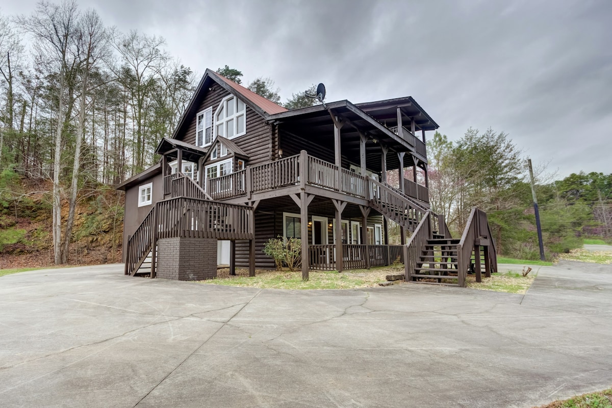 The mountain home is constructed of dark wood, featuring multiple levels and an expansive covered porch with ample seating. Surrounding trees provide a natural backdrop, while a large driveway offers parking space for several vehicles. The entrance is accessible via a set of stairs leading up to the porch.