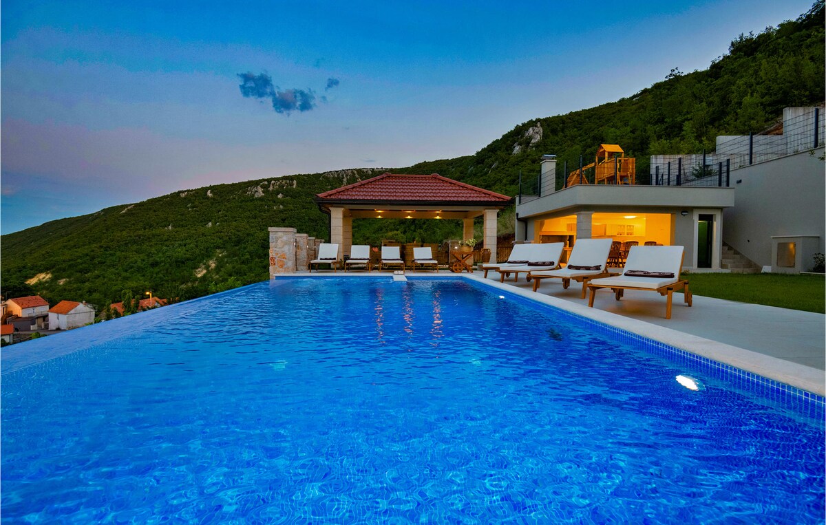 An inviting infinity pool is set against a backdrop of green hills, with several sun loungers positioned nearby. A shaded area with seating is visible in the distance, alongside the modern holiday home featuring expansive glass doors.