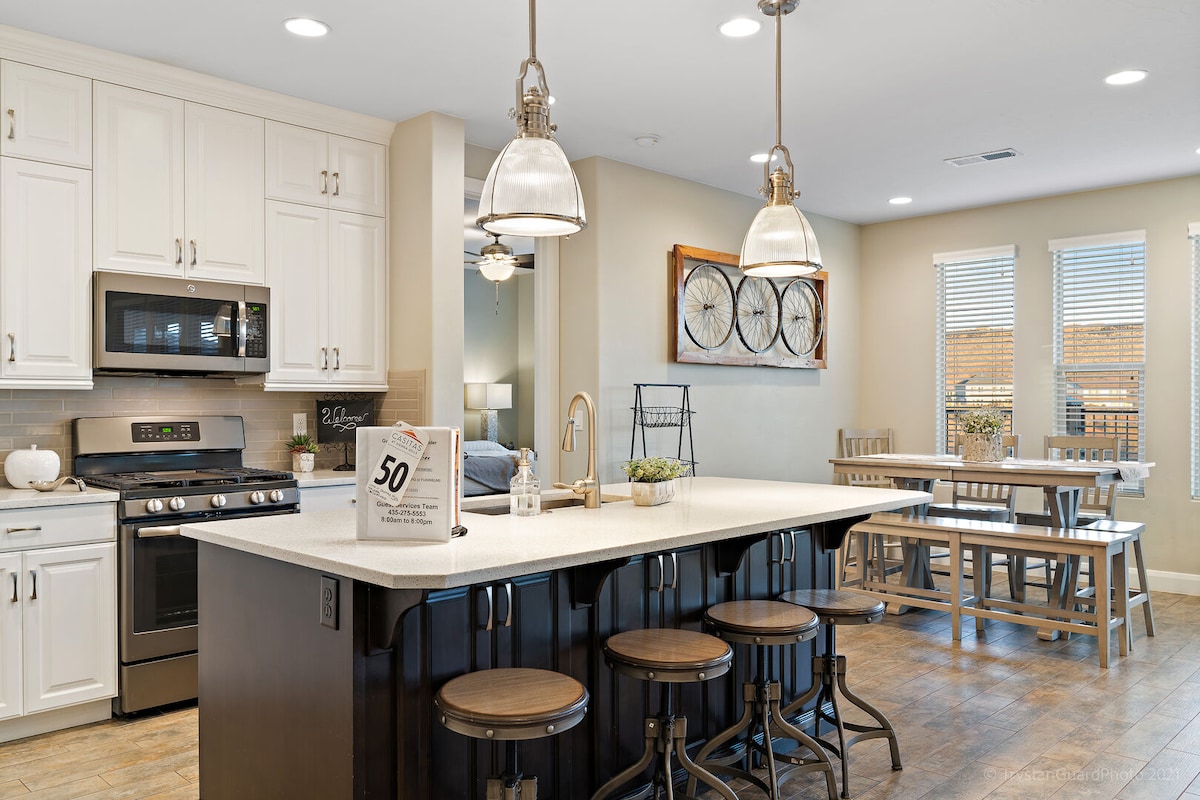 A modern kitchen features white cabinetry and stainless-steel appliances, including a microwave and stove. An island with seating for three contrasts with the dark cabinetry. A dining area is visible in the background, showcasing a wooden table surrounded by chairs and natural light from multiple windows.