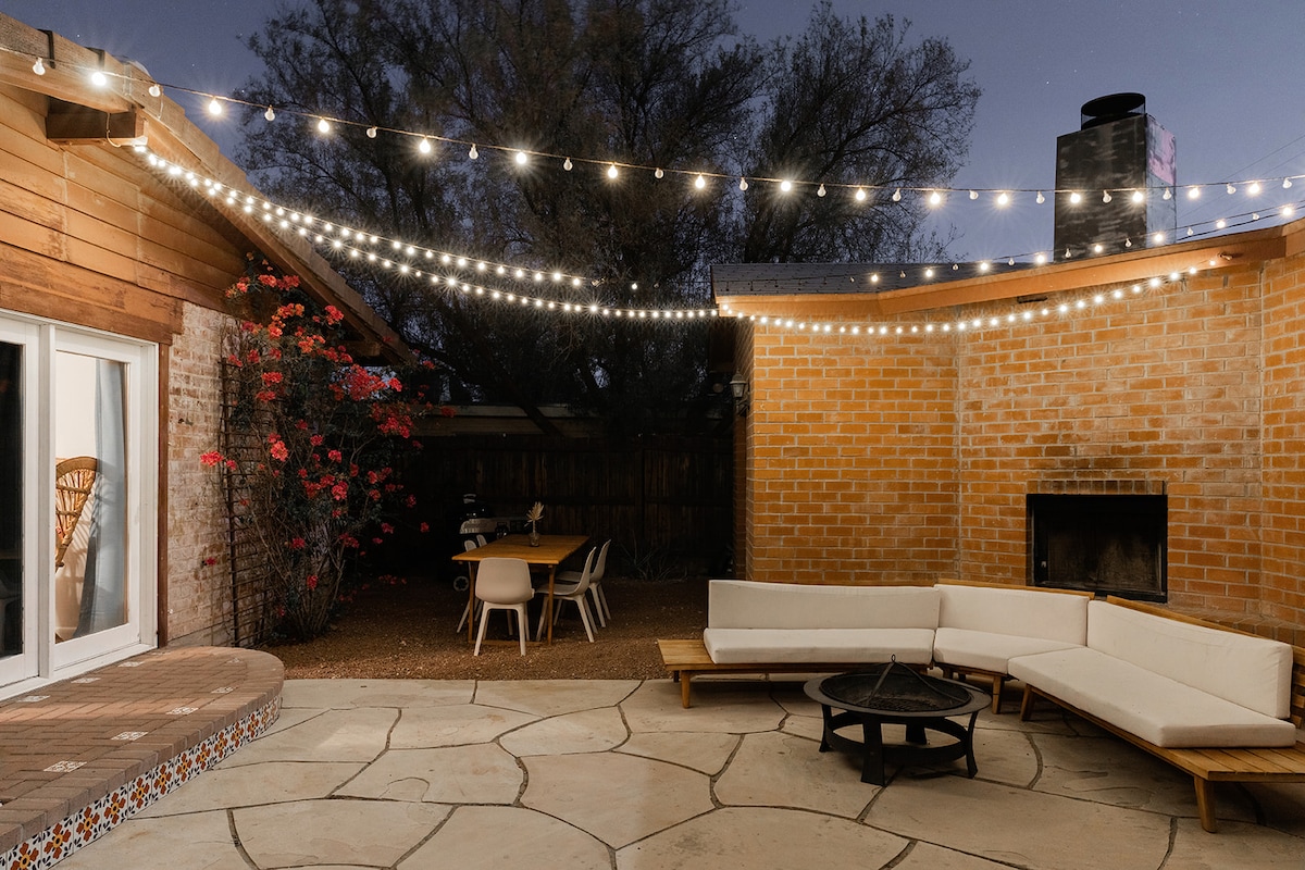 An outdoor patio area is illuminated by string lights, creating a warm ambiance during the evening. A circular fire pit is surrounded by a spacious sectional seating arrangement. A dining table is visible in the background, with blooming flowers adding a touch of color.