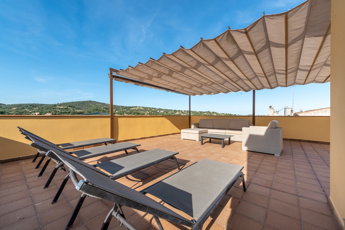 An upper terrace is presented with a shaded area and a comfortable seating arrangement. Several sun loungers are positioned on the tiled floor, providing a perfect space for relaxation. The backdrop features scenic views of rolling hills under a clear blue sky.