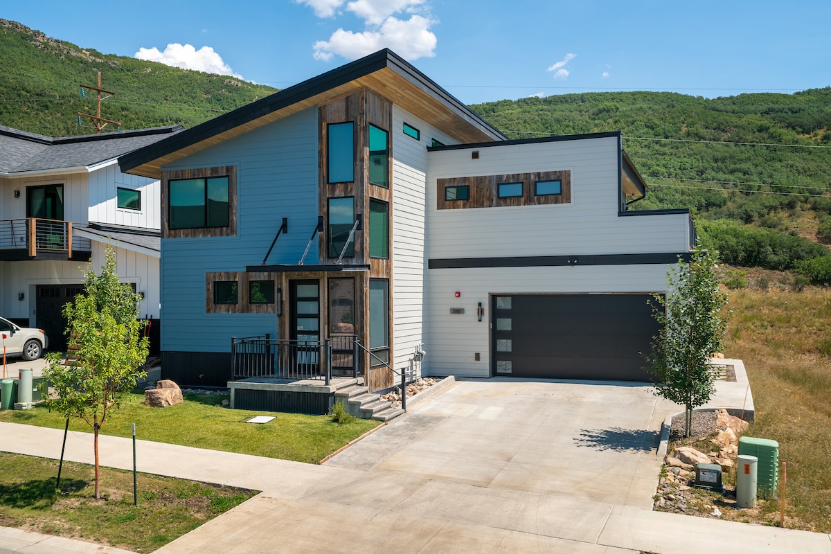 A modern two-story home is shown with a combination of wood and sleek siding. Large windows provide natural light, and the welcoming front porch features steps leading to the entrance. A two-car garage is positioned on the right, while grassy areas frame the property.