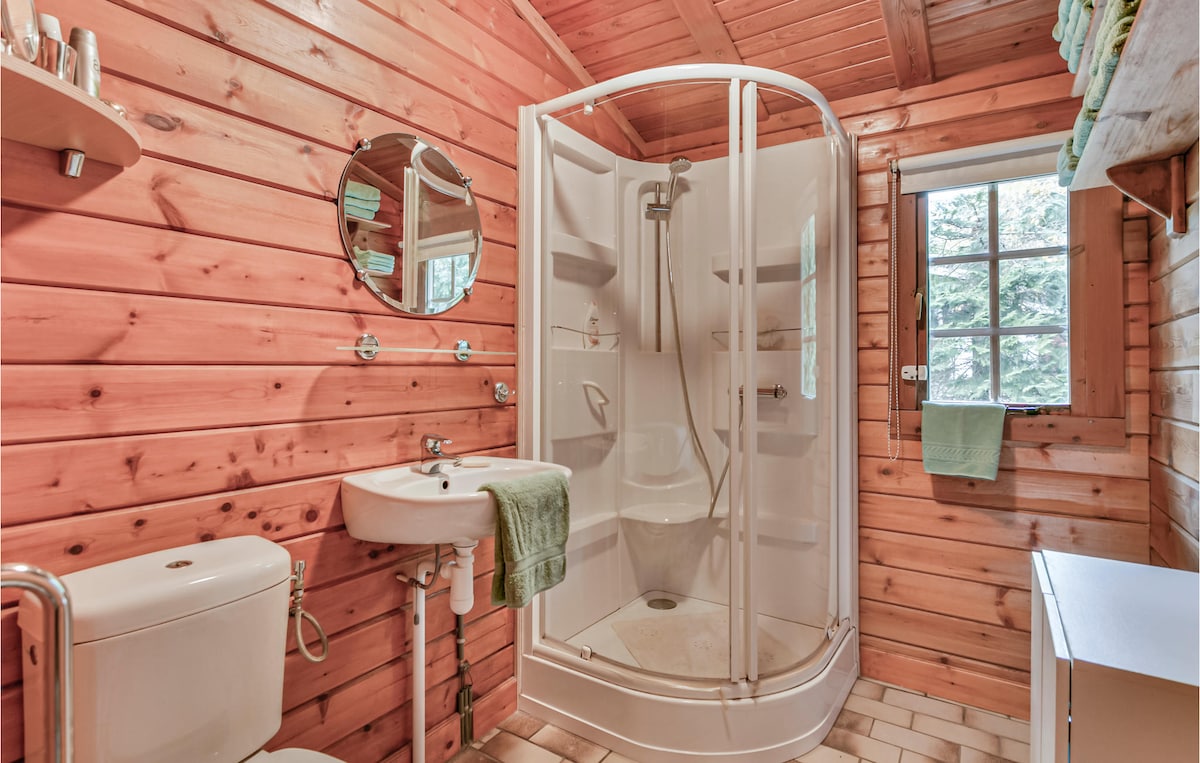 A bathroom features wooden wall paneling and a circular mirror above a small sink. A glass shower enclosure is present, and towels hang neatly nearby. A window allows natural light to enter, complementing the warm wood tones and greenery visible outside.