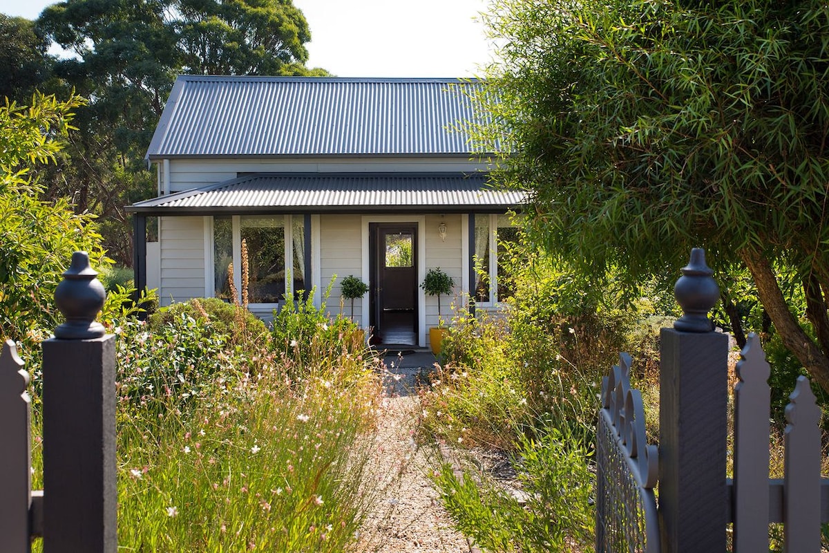 A charming country cottage with a metal roof is framed by lush greenery and flowering plants. A pathway leads to the entrance, where potted plants are placed on either side of the door, inviting visitors to enjoy the natural surroundings.