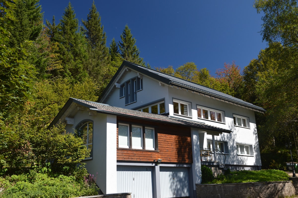 A two-story building is surrounded by lush greenery, showcasing a blend of white and brown exterior finishes. Multiple windows allow natural light to fill the interior, while a garage is visible at the base. The setting reflects a serene and natural environment.