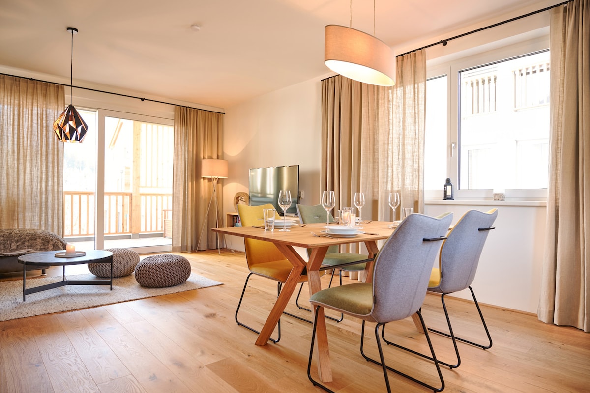 The open dining area features a wooden table set for four, surrounded by colorful chairs in soft shades. Natural light flows in through large windows with sheer curtains, illuminating the modern decor. A cozy seating arrangement with a round coffee table enhances the inviting atmosphere.