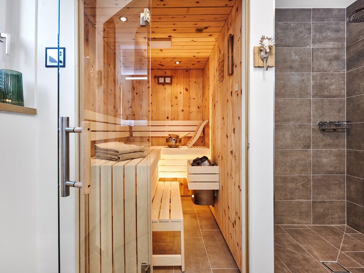 A private sauna is featured, constructed with warm wooden panels. The interior includes wooden benches and a small metal bucket, contributing to a relaxing environment. A clear glass door provides an unobstructed view of the sauna from the adjacent tiled shower area.