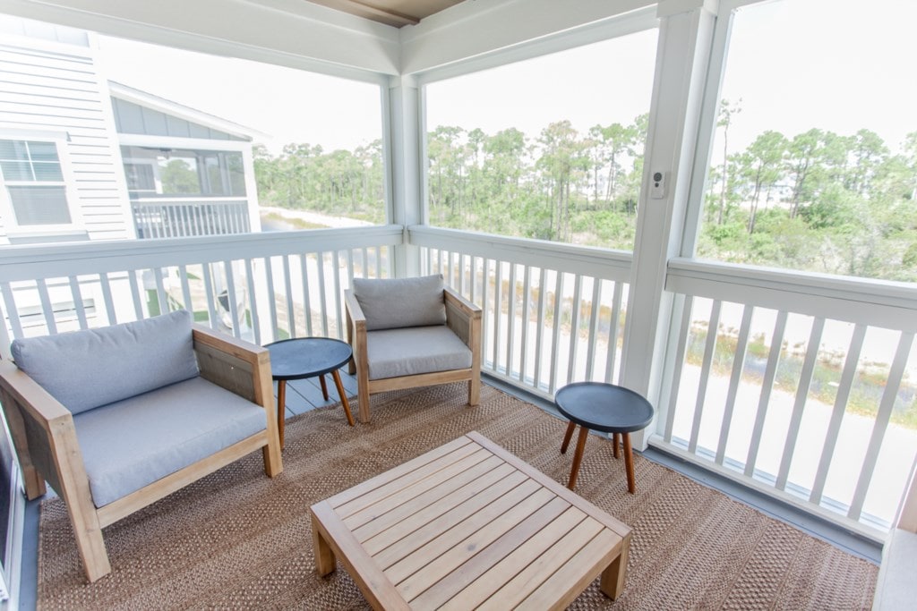 A spacious screened-in porch features two wooden chairs with soft cushions and a low wooden table. A natural fiber rug covers the floor, and the surrounding screens allow for a view of the trees outside, creating a serene and inviting space.