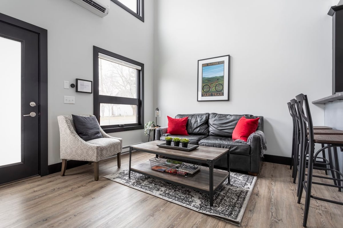 A modern living area features a dark leather sofa adorned with red cushions, complemented by a cozy armchair. A low coffee table holds decorative plants, while a large window enhances natural lighting. A stylish rug adds texture to the wooden floor.